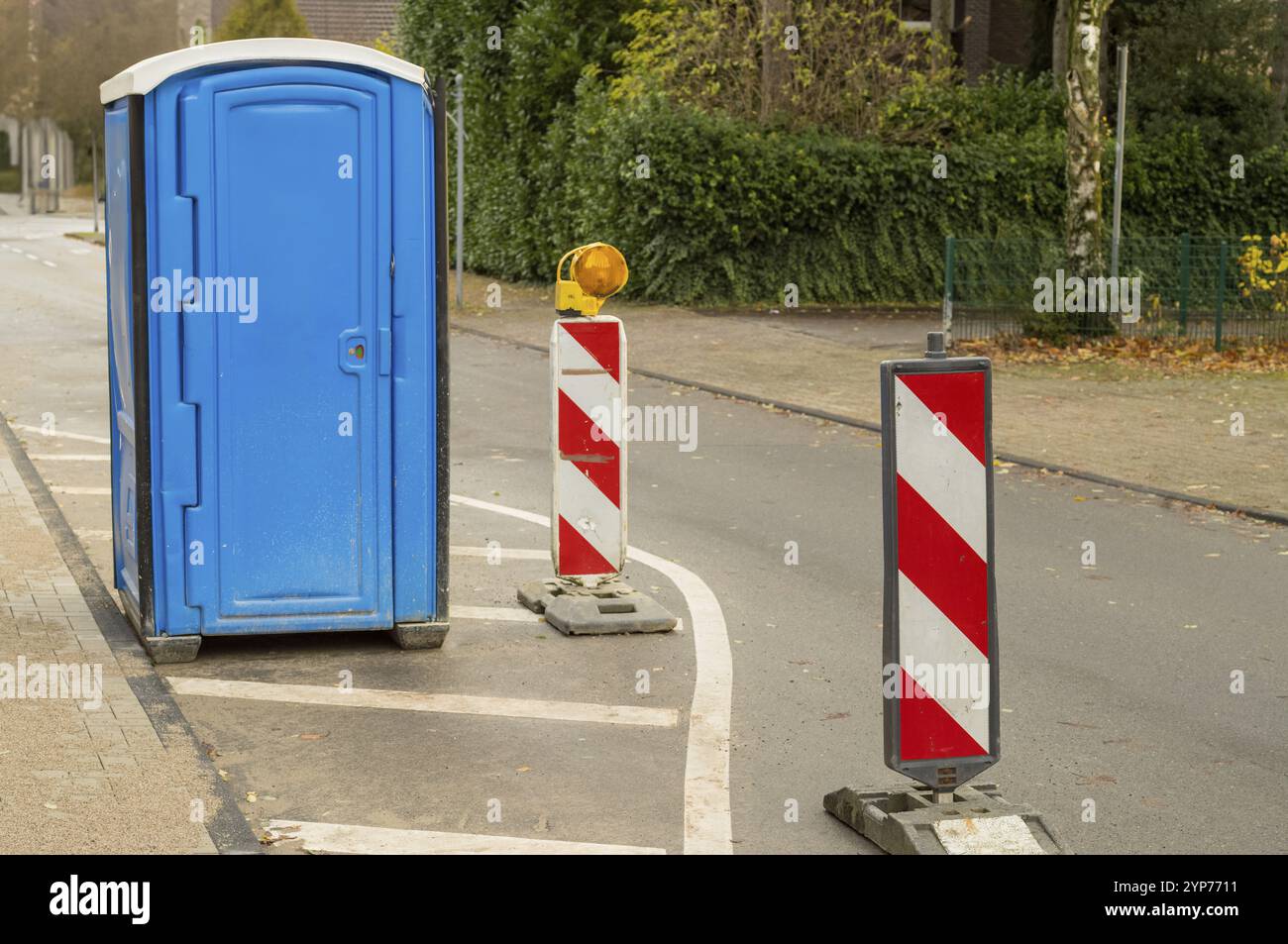 La toilette portatile blu si trova su una strada accanto ai pali di segnalazione del traffico che indicano i lavori stradali Foto Stock