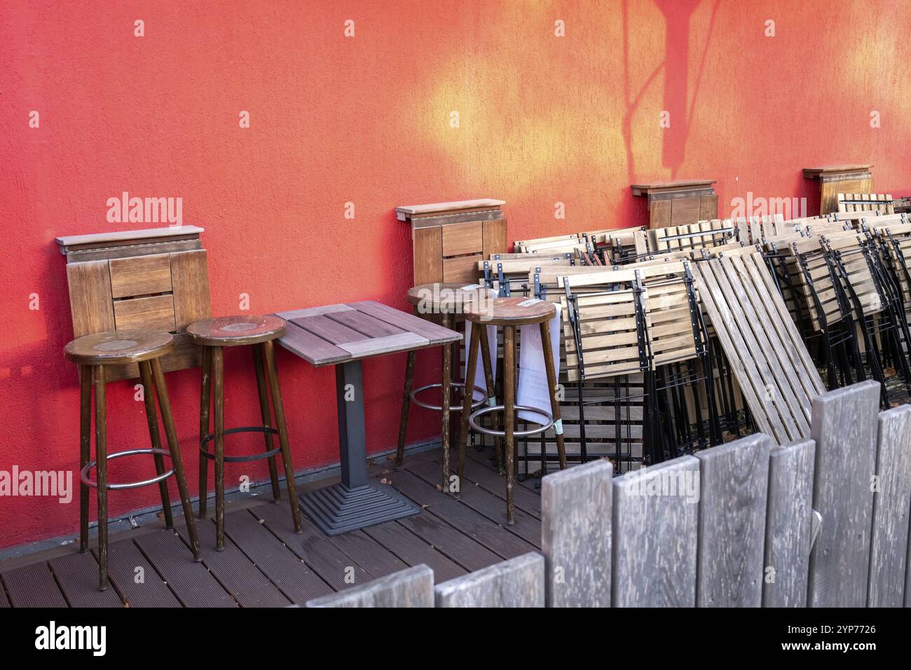 Area esterna con sgabelli in legno e un tavolo contro una vivace parete rossa Foto Stock