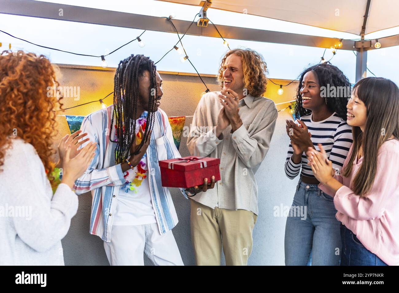 Uomo africano che ringrazia i suoi amici multietnici per il regalo di compleanno che gli hanno dato durante una festa in terrazza Foto Stock