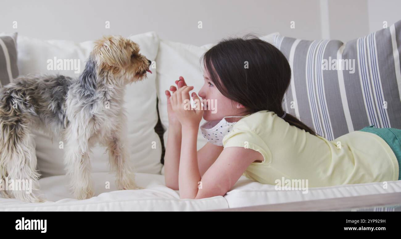 Ragazza caucasica che trascorre il tempo a casa, sdraiata sul divano con maschera facciale, giocando con il cane da compagnia, in slow mo Foto Stock