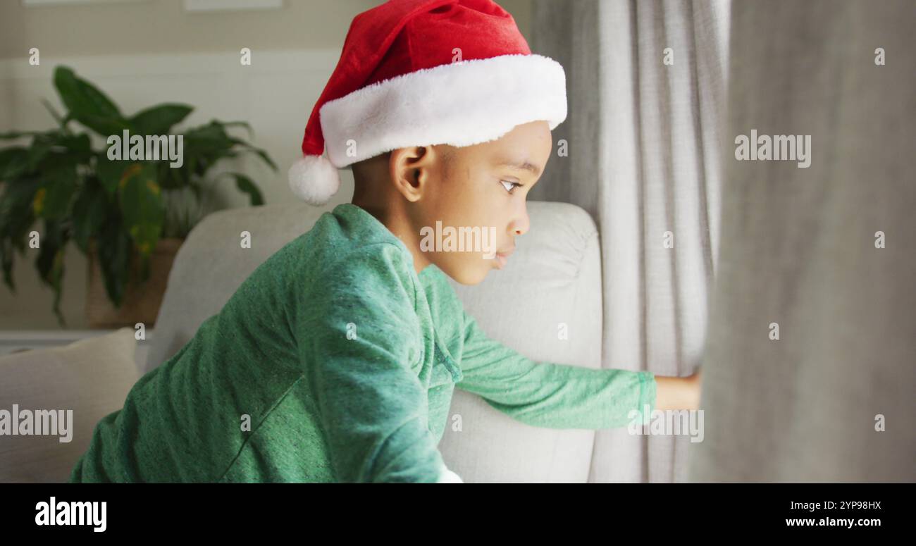 Triste ragazzo afroamericano che indossa il cappello di babbo natale guardando fuori dalla finestra nel periodo natalizio Foto Stock