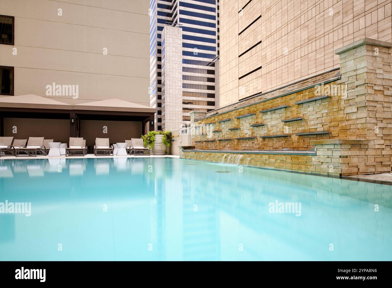 Una tranquilla piscina dell'hotel presenta una rilassante parete d'acqua e sedie a sdraio, sullo sfondo di eleganti grattacieli in una giornata di sole, perfetta per rilassarsi Foto Stock