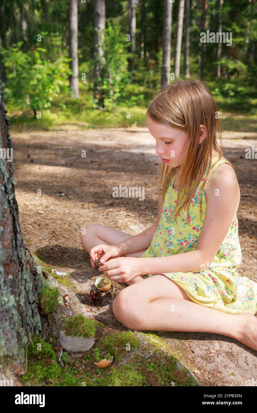 Una giovane ragazza con un abito floreale giallo si siede in una foresta illuminata dal sole, creando una scena in miniatura alla base di un tronco d'albero ricoperto di muschio Foto Stock