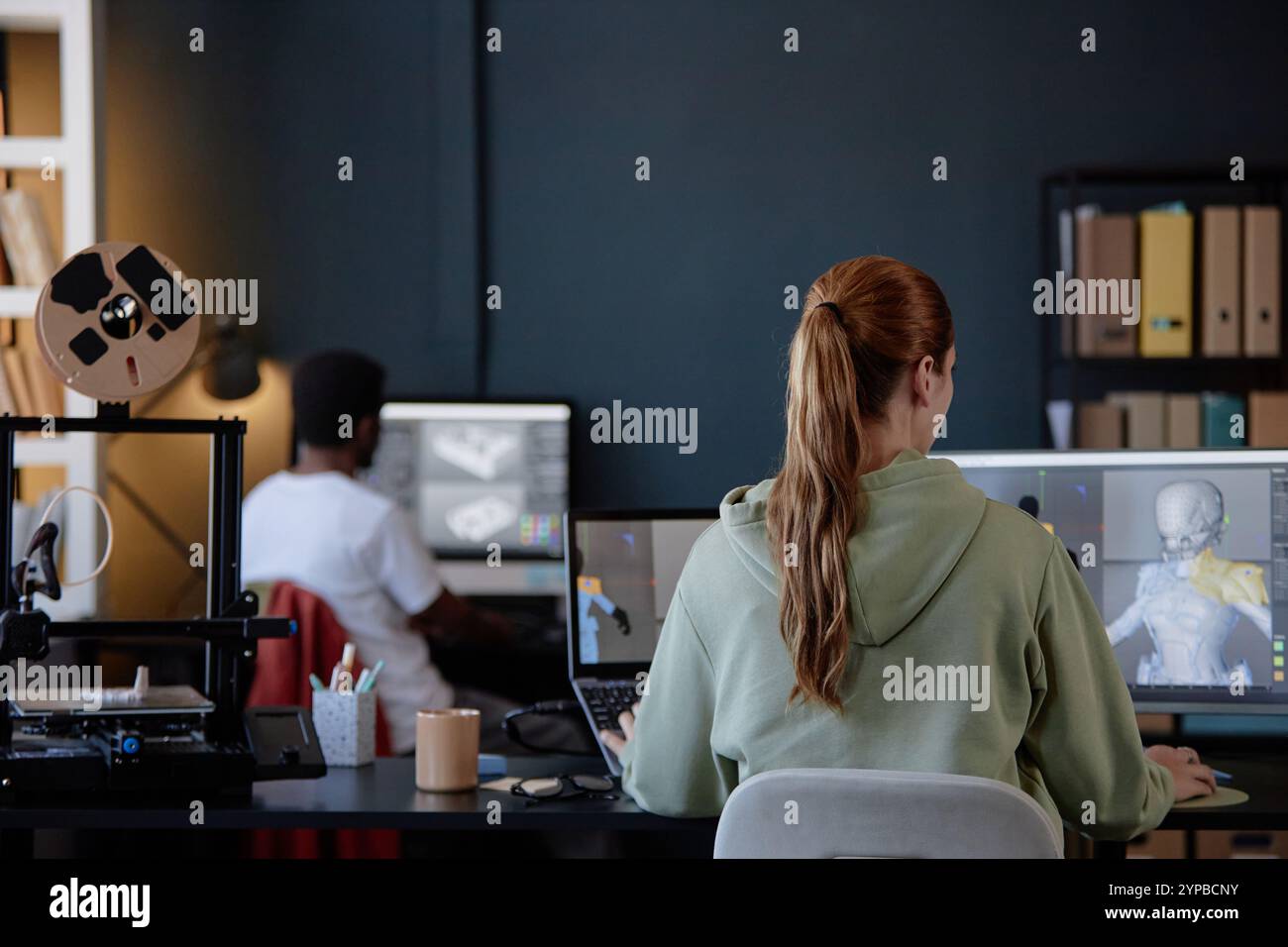 Persone coinvolte nella codifica e nella progettazione di modelli 3D in un ambiente di ufficio moderno con vari gadget e strumenti. Ripiani pieni di file e cartelle in background Foto Stock