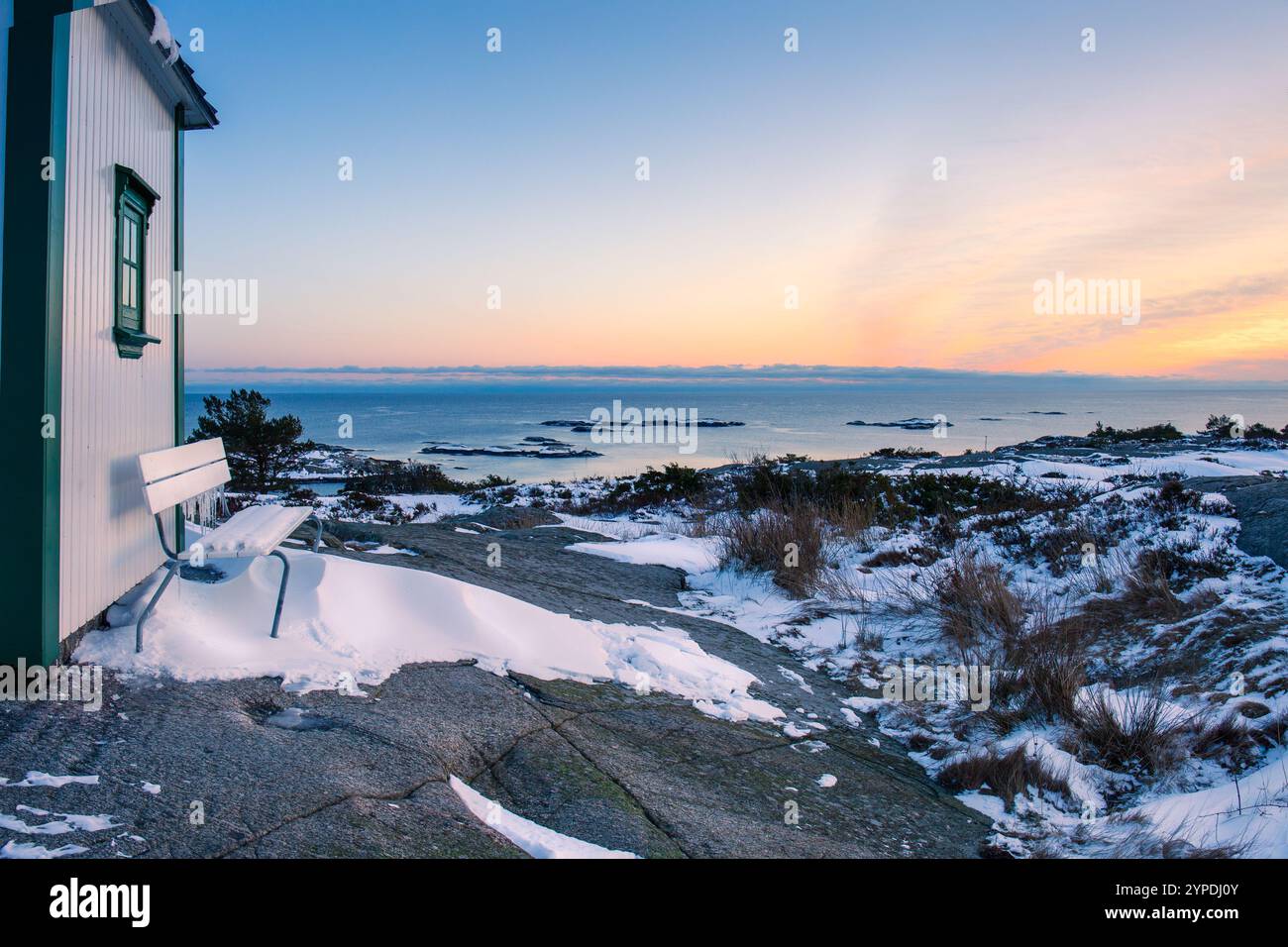 Casa in legno bianco con dettagli verdi accanto a una panchina coperta di neve che si affaccia su un vasto paesaggio marino invernale al tramonto. Caldo, tranquillo e adatto al mare Foto Stock