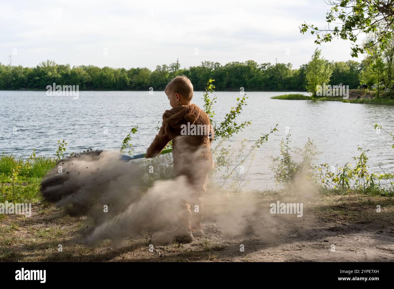 Ragazzo che gioca vicino al lago, creando una spettacolare nuvola di polvere nell'aria con la pala nelle giornate di sole in un ambiente naturale. Divertimento d'infanzia, attività all'aperto Foto Stock