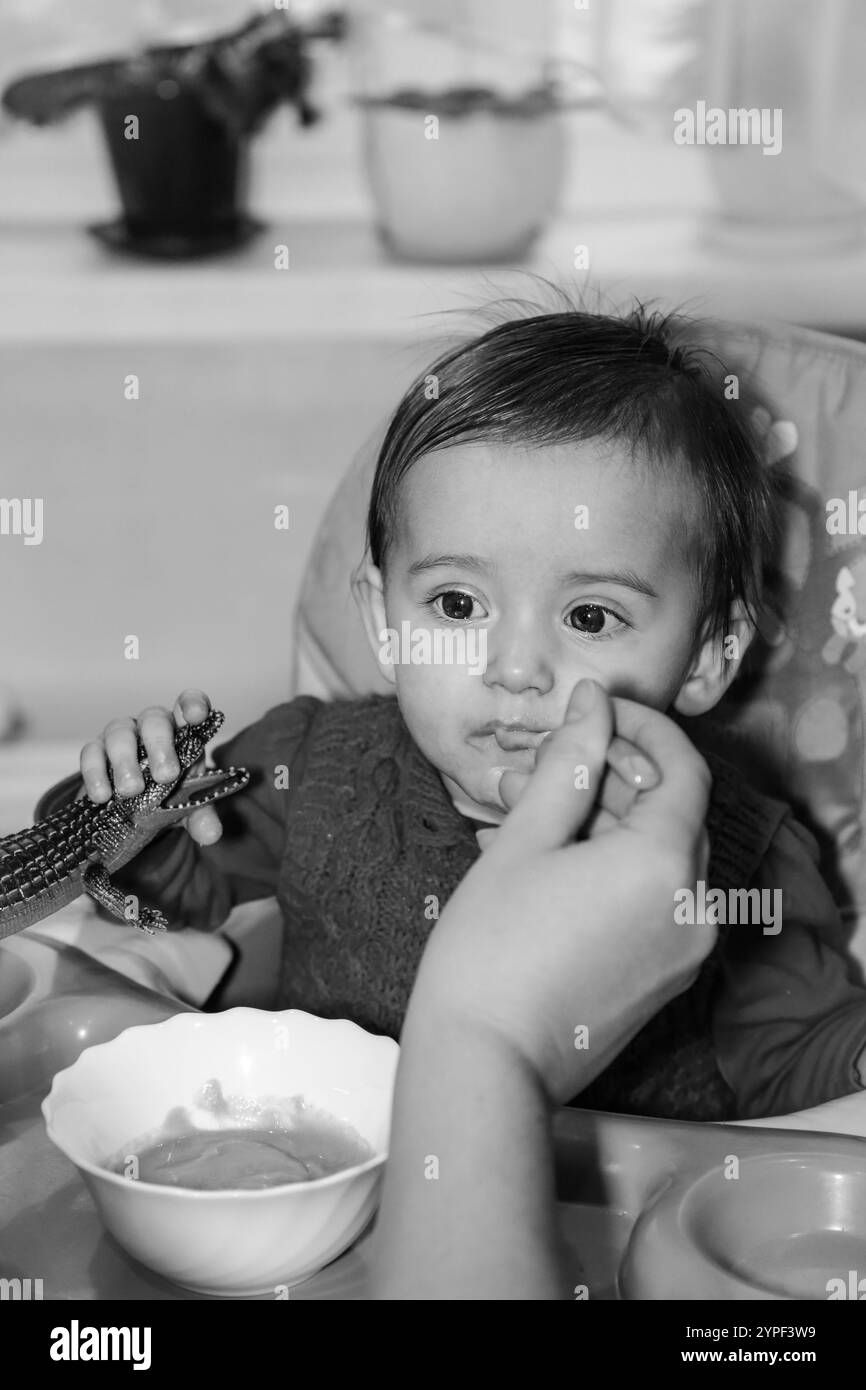 Un bambino ama mangiare nutriente quando viene nutrito al cucchiaio mentre è seduto a un tavolo in cucina, foto monocromatica Foto Stock
