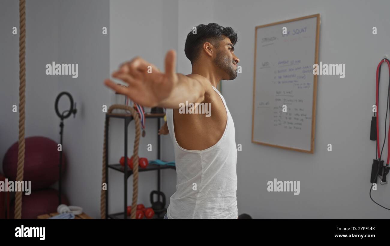 Giovane che si allunga le braccia in una palestra coperta circondata da attrezzature fitness e tabelle di allenamento, dimostrando una particolare attenzione al fitness fisico e alla guarigione Foto Stock