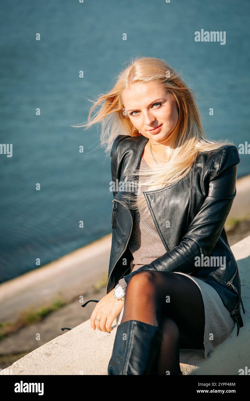 Giovane donna bionda seduta sul fiume con giacca in pelle, stivali e collant, godendosi una giornata di sole Foto Stock