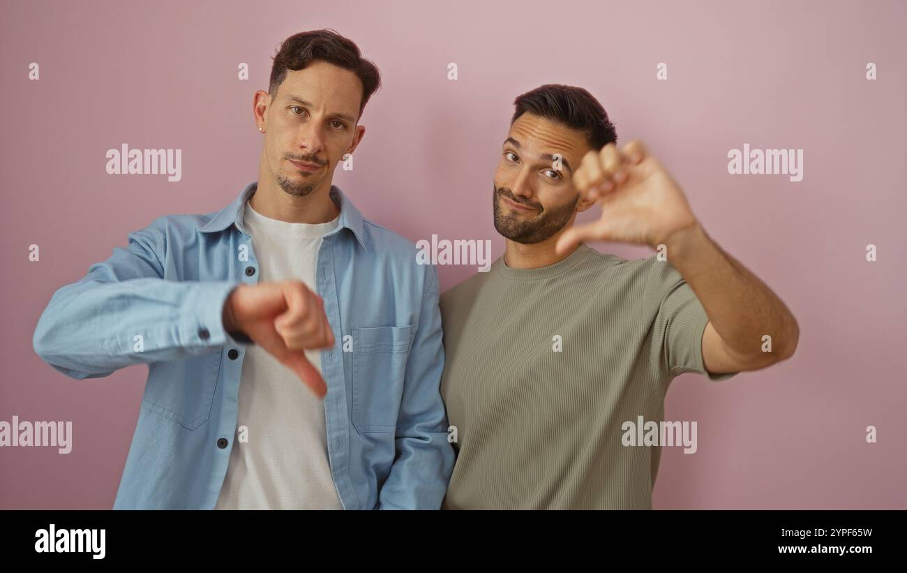 Coppia gay in piedi insieme facendo gesti con il pollice giù su uno sfondo rosa, abbracciando la loro rappresentazione lgbtq con abbigliamento casual e viso Foto Stock