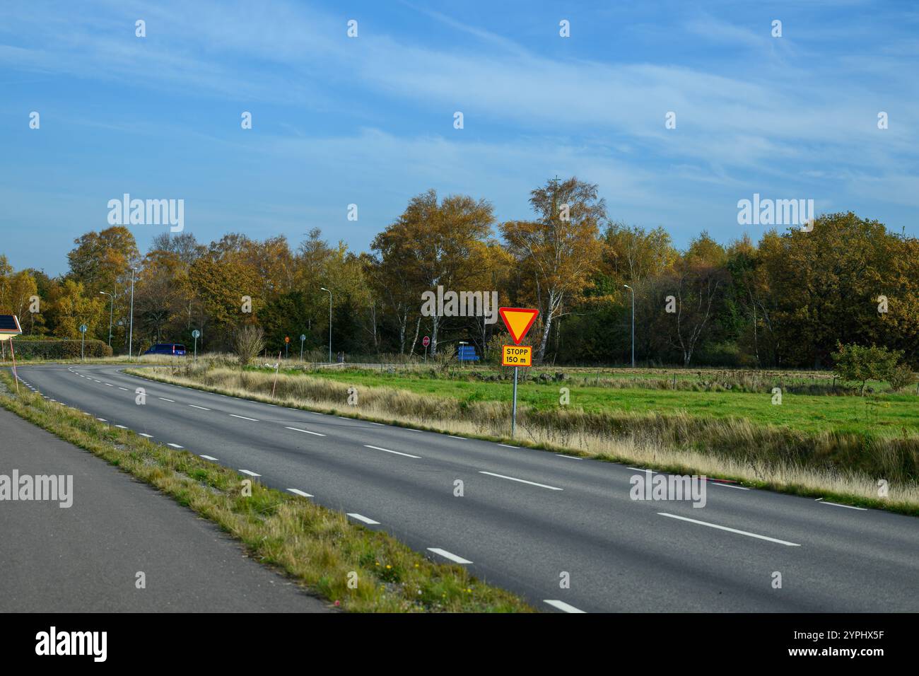 Una strada rurale si estende in lontananza, incorniciata da vibranti alberi autunnali su entrambi i lati. Un segno di resa viene visualizzato in modo prominente per indicare la necessità di Foto Stock