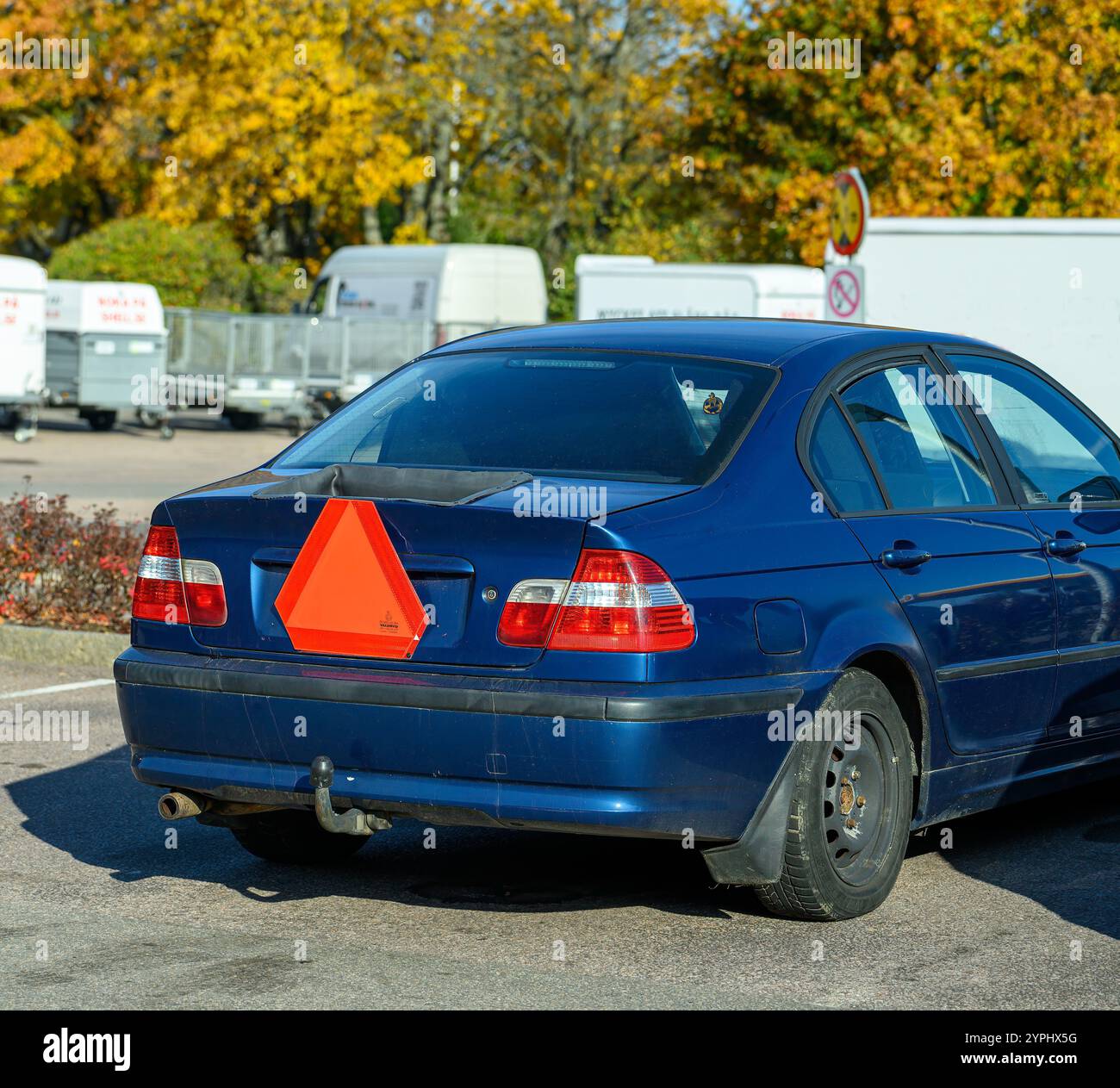 Un trattore di colore blu è parcheggiato molto, con una caratteristica forma triangolare arancione sul retro. Gli alberi circostanti mostrano vivaci colori autunnali sotto Foto Stock
