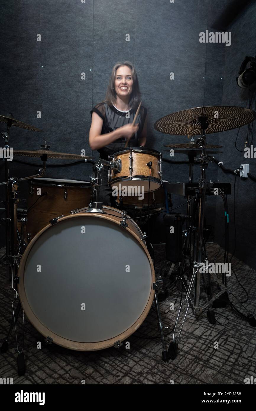 Batterista sorridente che si esibisce sul set della batteria in uno studio insonorizzato professionale. La scena vibrante enfatizza l'energia, la creatività e la gioia della creazione di musica dal vivo e dell'immagine verticale Foto Stock