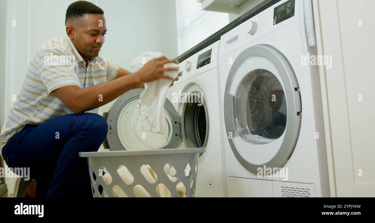 Vista laterale dell'uomo nero medio adulto che lava i vestiti in lavatrice in una casa confortevole Foto Stock