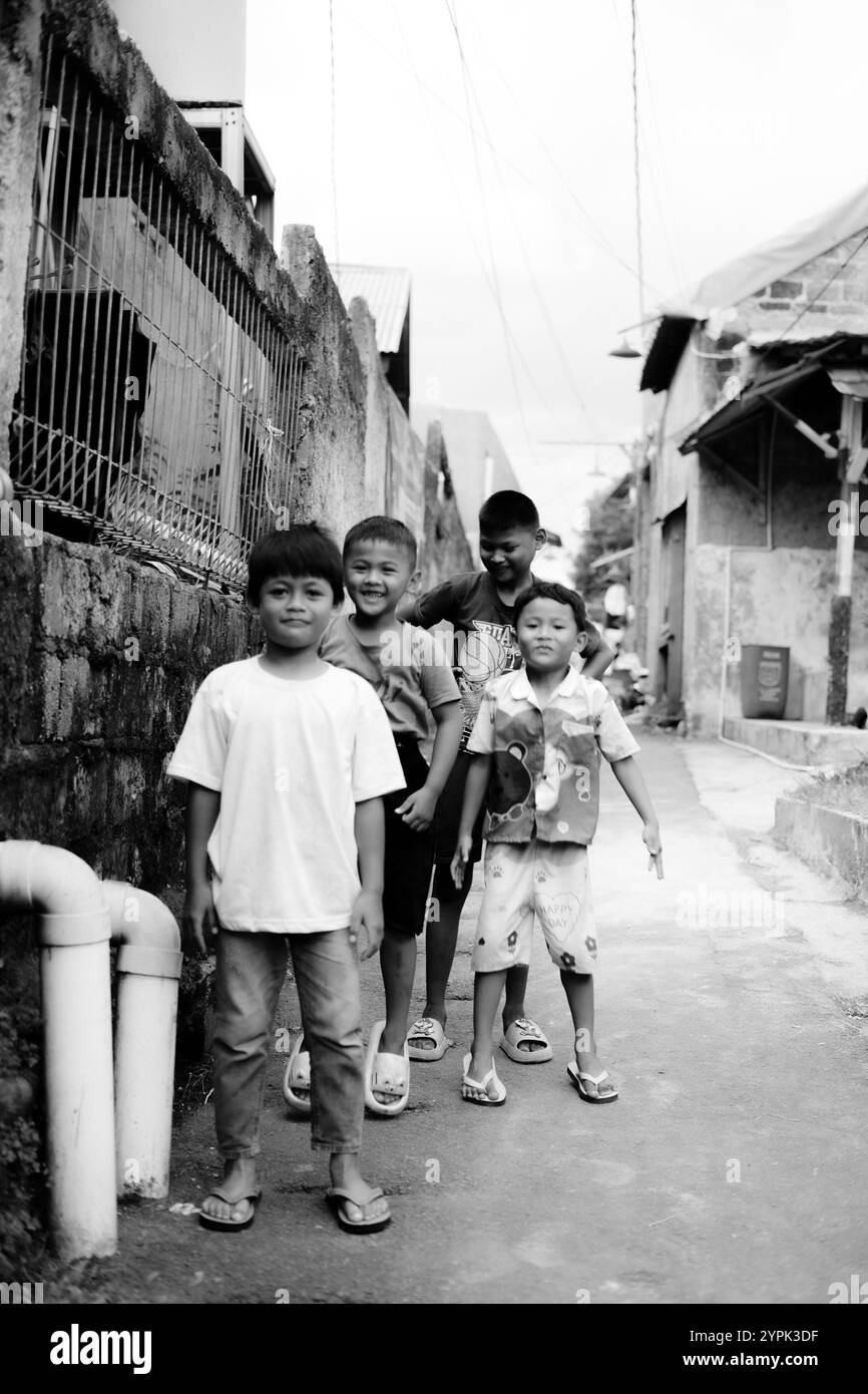 Bogor, 24 novembre 2024. Quattro bambini piccoli in piedi insieme in un vicolo stretto, sorridenti e guardano intorno in un vivace ambiente del quartiere Foto Stock