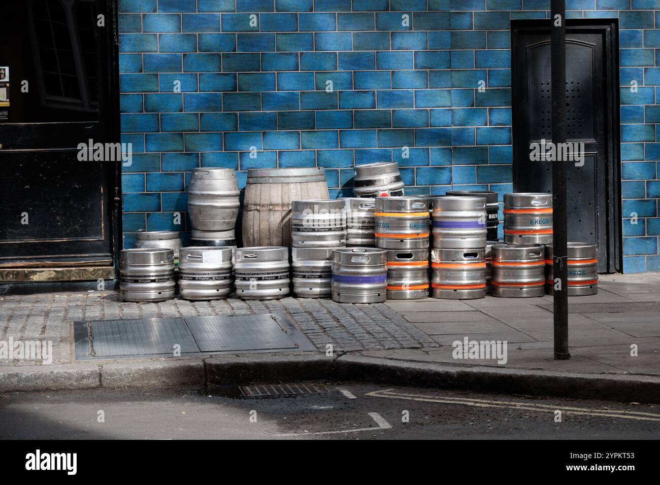 Una pila di fusti di birra in metallo contro pareti di piastrelle blu fuori dal pub di Londra, che mostrano un contrasto tra botti di argento industriali e vibrante sfondo urbano Foto Stock