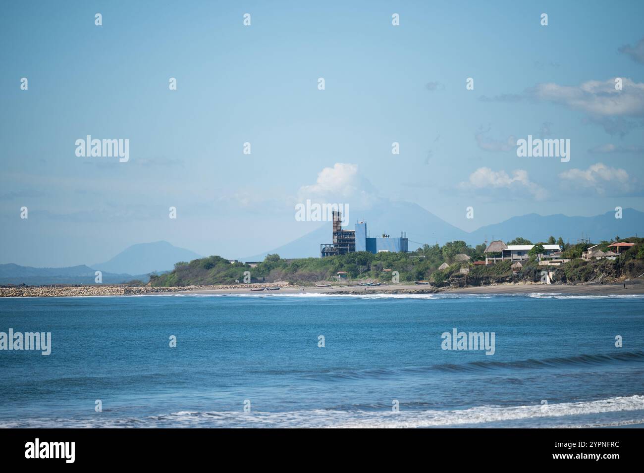 Litorale con onde dolci e strutture industriali visibili sullo sfondo sotto il cielo soleggiato. Foto Stock