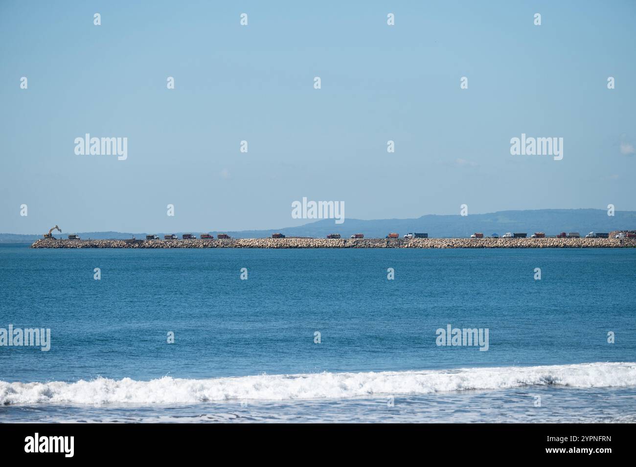 Il lungo molo roccioso si estende nell'oceano azzurro sotto il cielo limpido, con le onde che si infrangono alla sua base. Foto Stock