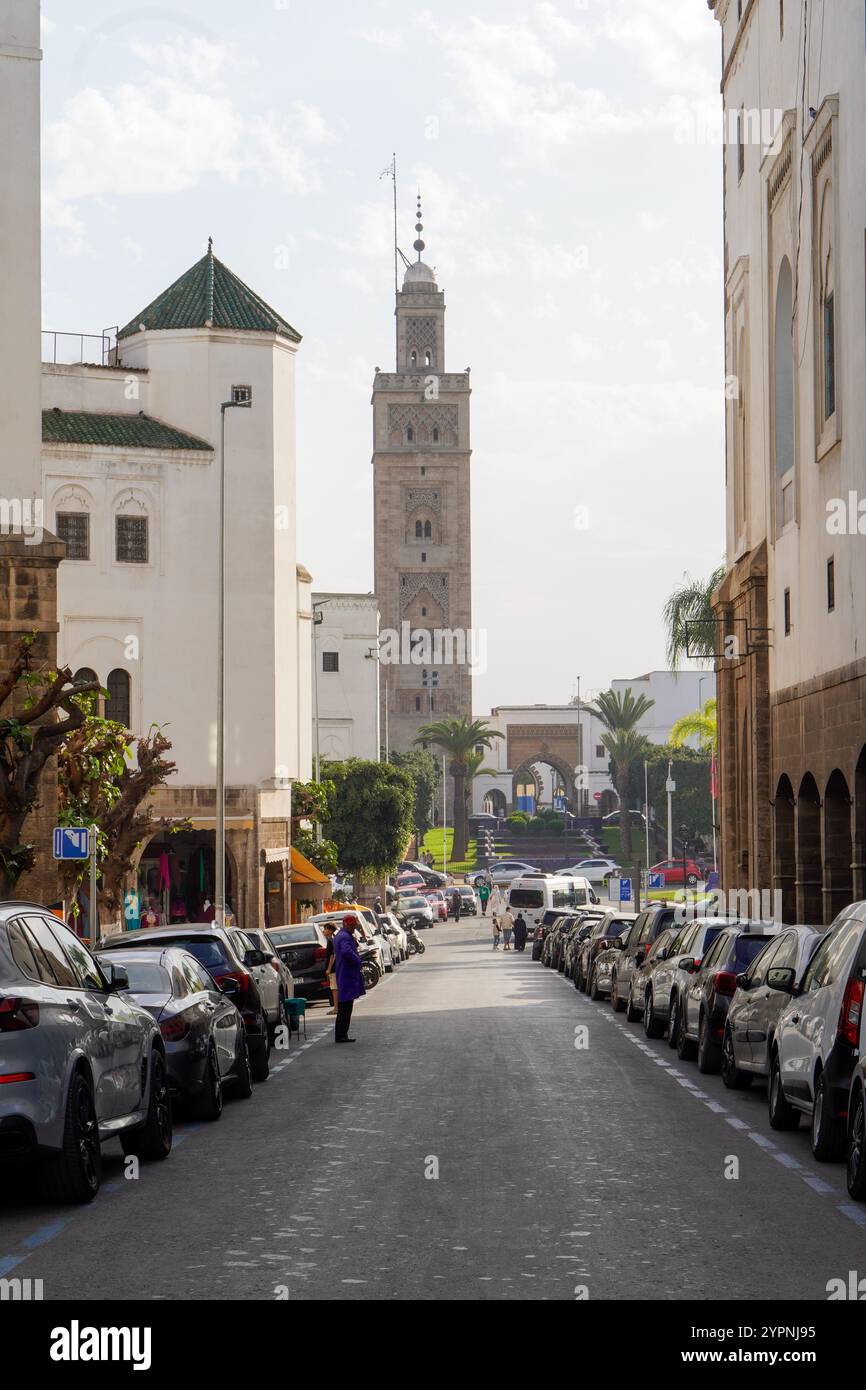 Vivace strada nel quartiere Habous di Casablanca, in Marocco, con auto parcheggiate su entrambi i lati e un'importante moschea con un alto minareto sul retro Foto Stock