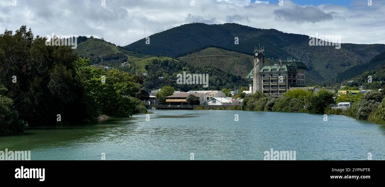 Una tranquilla vista sul fiume con vegetazione lussureggiante, un caratteristico edificio a torre dell'orologio e colline boscose sullo sfondo sotto un cielo parzialmente nuvoloso. Foto Stock