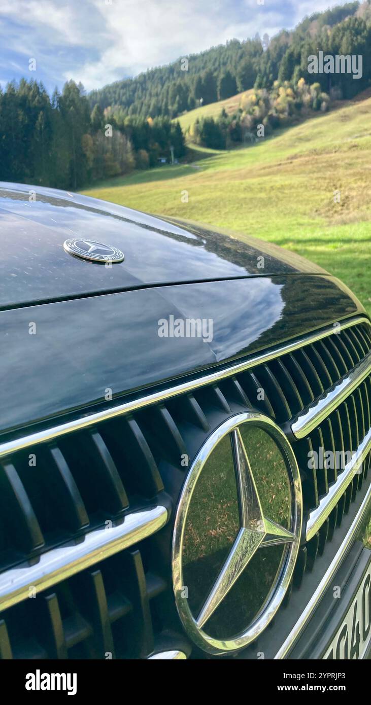 Parte di una griglia e cappuccio di una Daimler Mercedes Benz Classe e nera con pendenze delle alpi tedesche sullo sfondo - Immagine stock catturata con smartphone