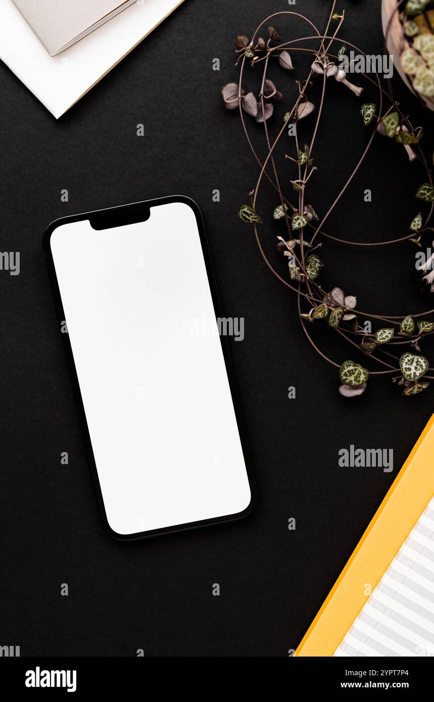 Smartphone moderno su sfondo nero da tavolo con delicati decori di piante domestiche, blocchi appunti gialli e grigi - elegante configurazione dello spazio di lavoro. Vista dall'alto. Foto Stock
