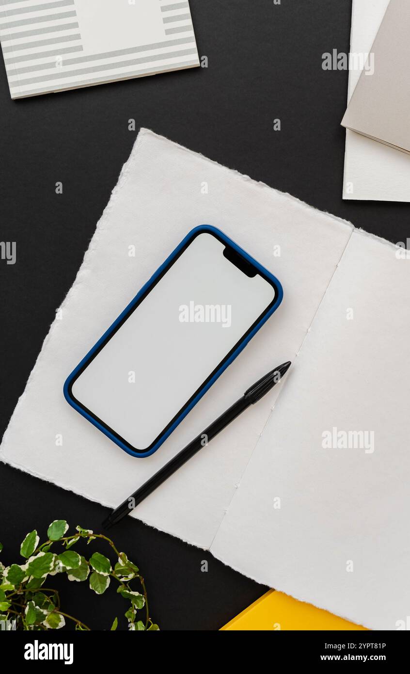 Moderno smartphone con custodia blu su blocco note aperto su sfondo nero del tavolo con decorazione verde della pianta domestica e altri notebook - elegante spazio di lavoro Foto Stock