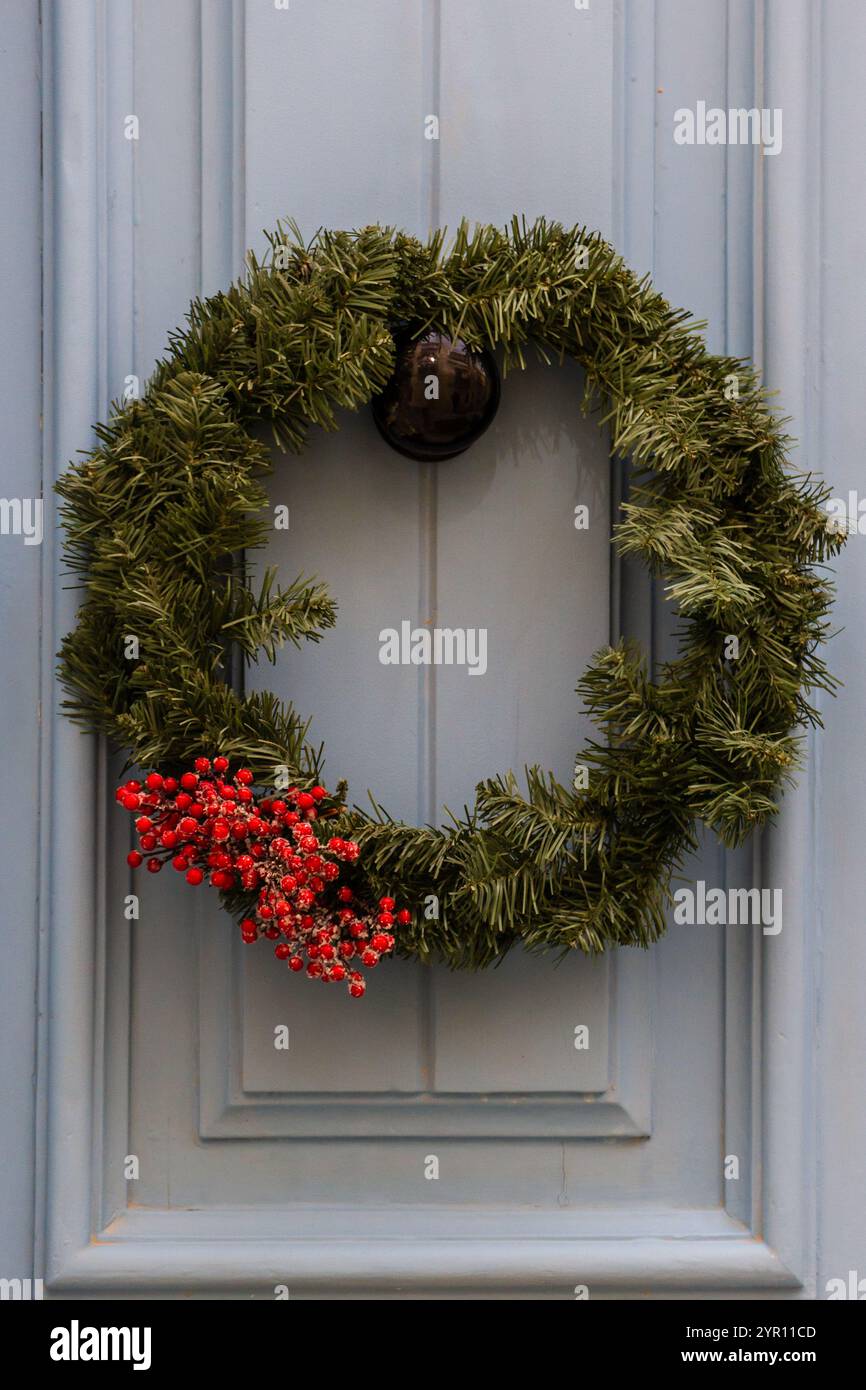 Una classica corona di Natale sempreverde adornata da vivaci frutti di bosco rossi è appesa su una porta blu chiaro, evocando spirito natalizio e calore. Foto Stock