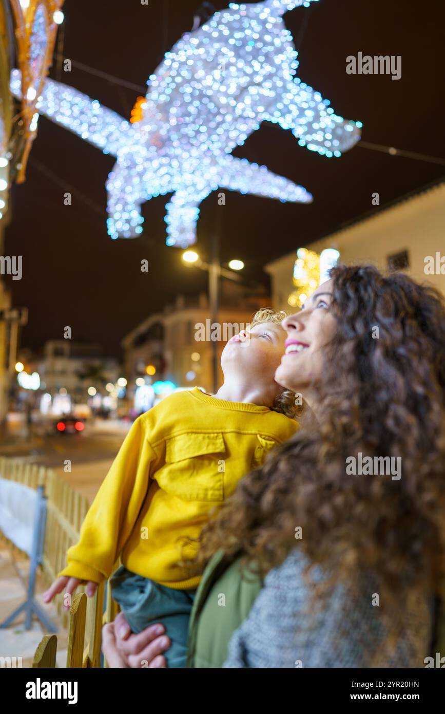 Madre e figlio ammirano le luci di natale in città di notte, sentendo magia e gioia durante le festività natalizie Foto Stock