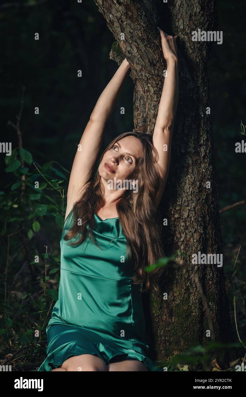 Donna sorridente che si riposa vicino all'albero con corteccia ruvida, guarda in alto e abbraccia il tronco. Vestito verde, capelli lunghi Foto Stock