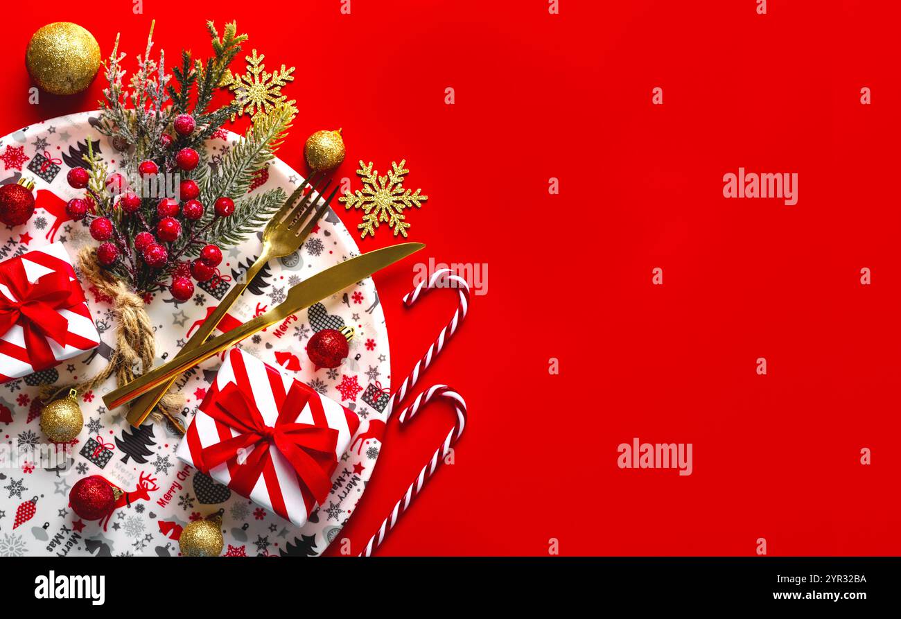 Cena di Natale. Vista dall'alto delle posate dorate e degli ornamenti natalizi su un piatto bianco con motivi natalizi con spazio per il testo su un dorso rosso Foto Stock