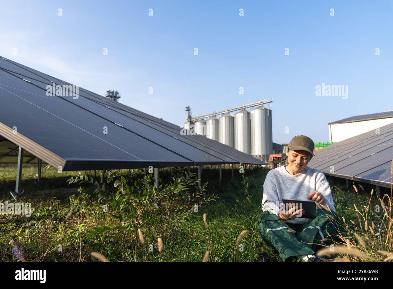 Agricoltore donna con tablet digitale in una moderna azienda agricola che utilizza pannelli solari. Silos agricoli sullo sfondo. Foto Stock