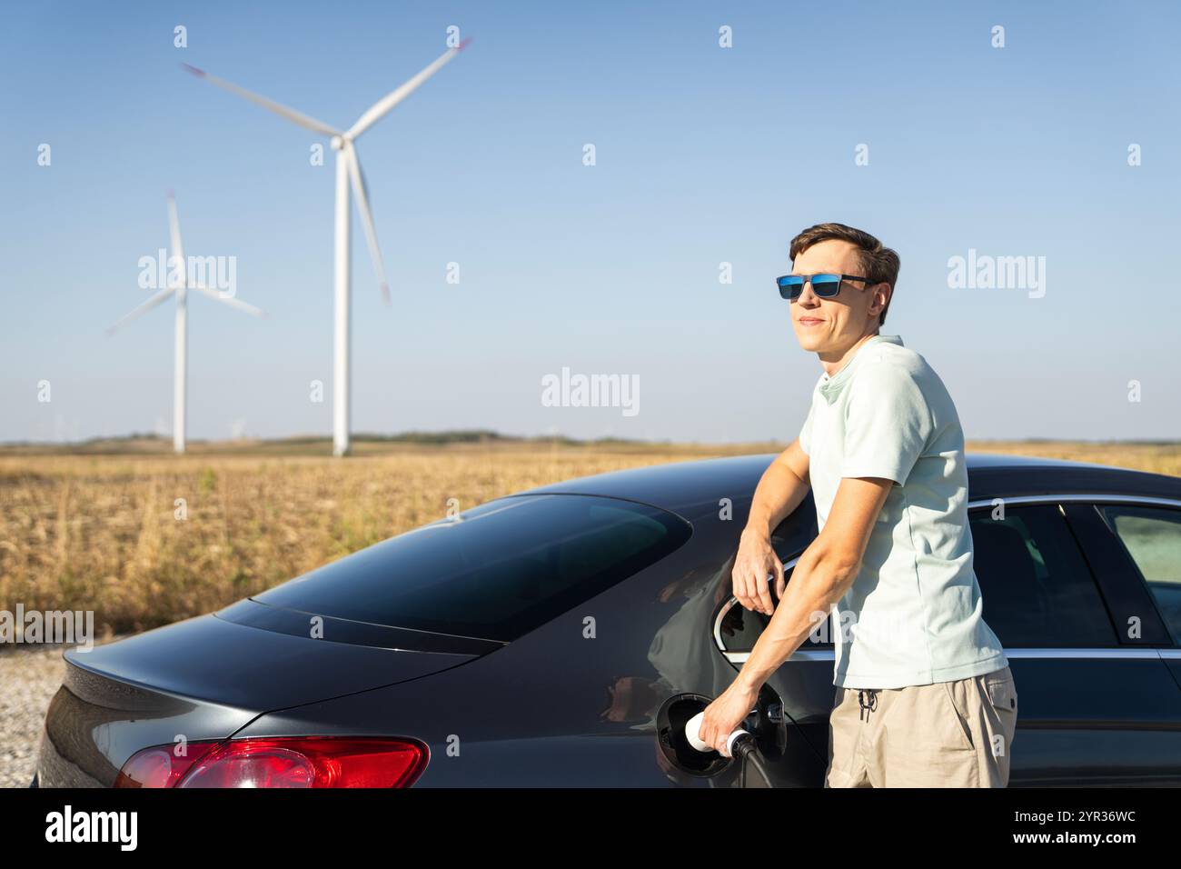 Uomo con auto elettrica in carica. Turbine eoliche sullo sfondo. Foto Stock