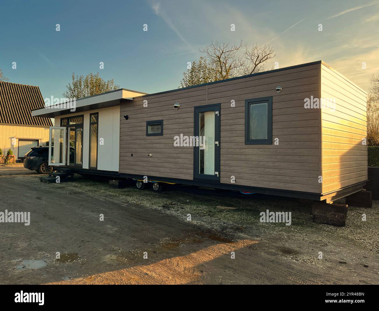 Una moderna casa mobile con un esterno in legno, rialzata su blocchi, situata all'aperto in un cantiere sotto un caldo cielo al tramonto, con un ripostiglio. Foto Stock