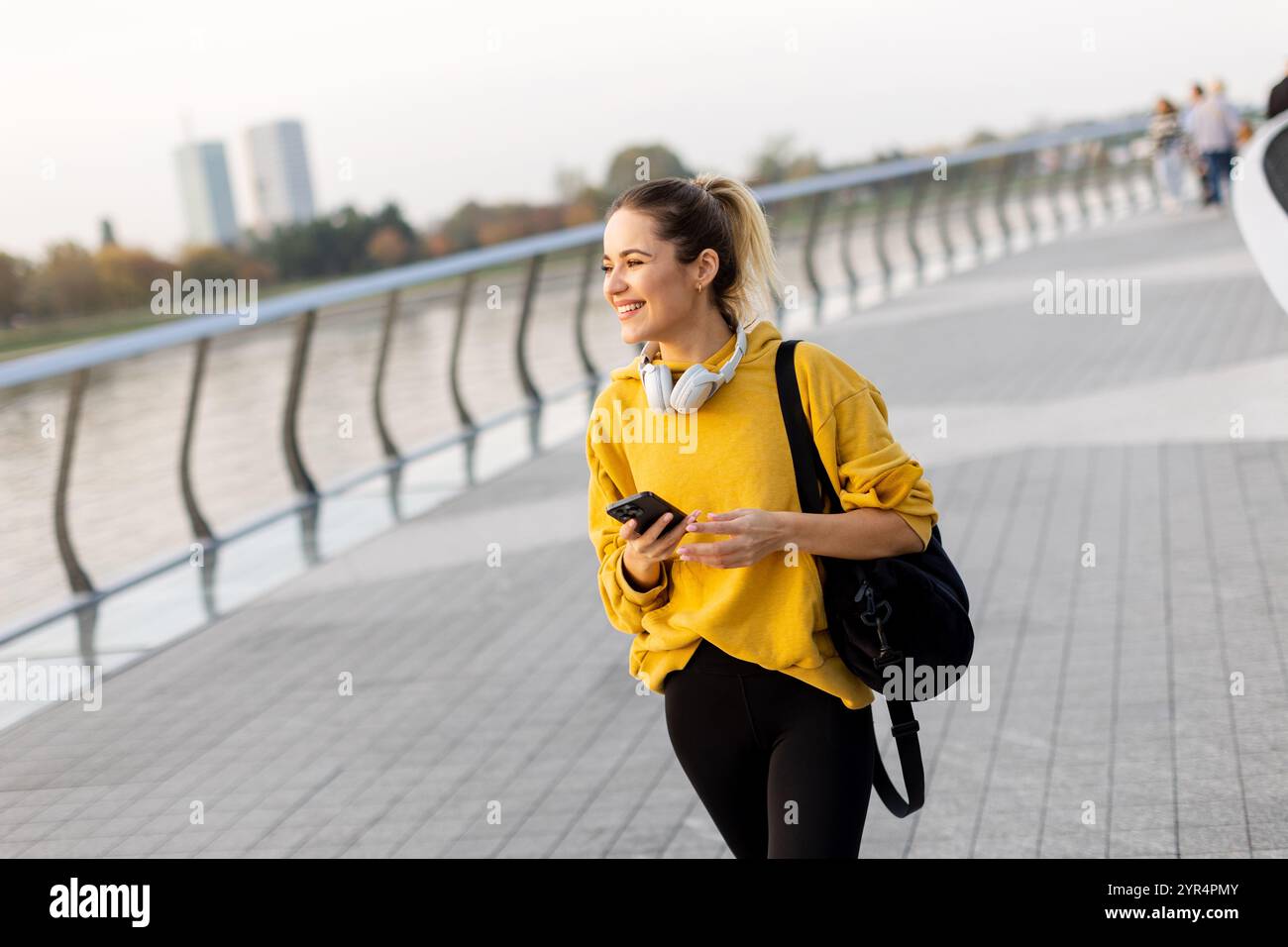 Una giovane donna sorridente cammina lungo un sentiero lungo il fiume in autunno, vestita con un comodo maglione giallo e cuffie. L'atmosfera serena cattura la sua gioia Foto Stock