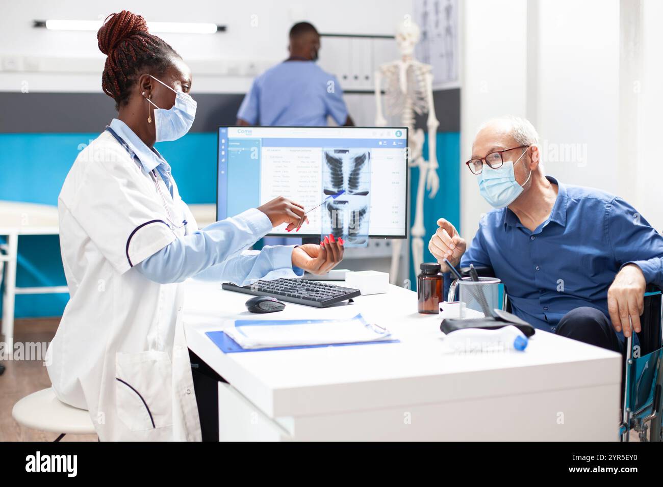 Paziente in pensione caucasica in consultazione con un medico donna, discutendo i risultati dei raggi x. Il medico afroamericano mostra l'immagine della radiografia polmonare all'uomo anziano in sedia a rotelle e spiega la diagnosi. Foto Stock