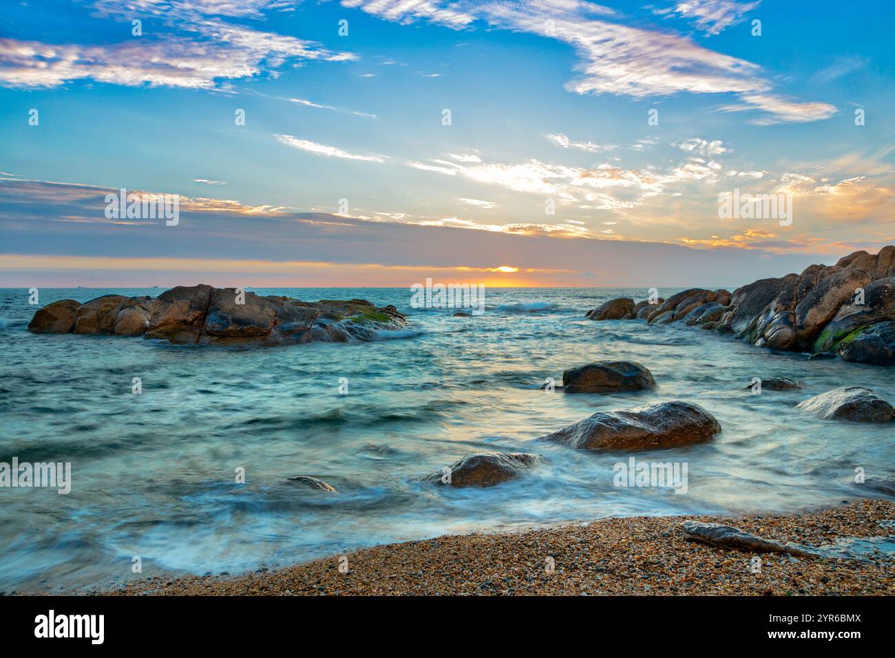 La luce soffusa del sole bagna una tranquilla scena oceanica, evidenziando rocce e una spiaggia di sabbia in primo piano, creando una tranquilla e pittoresca landsca costiera Foto Stock