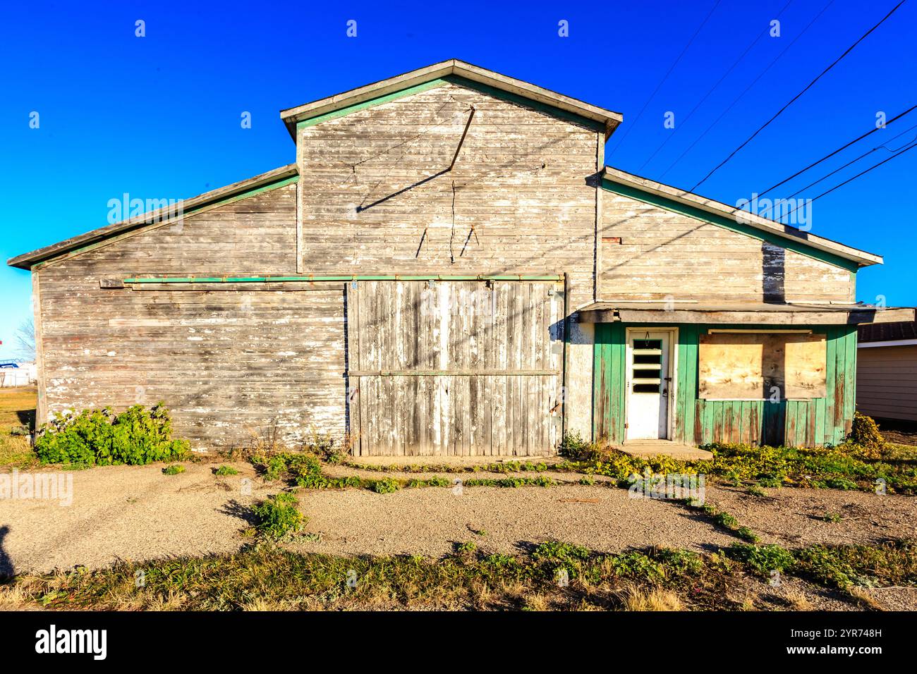 Un grande e vecchio edificio con una porta verde e finiture bianche. L'edificio è circondato da molte erbacce e erba Foto Stock