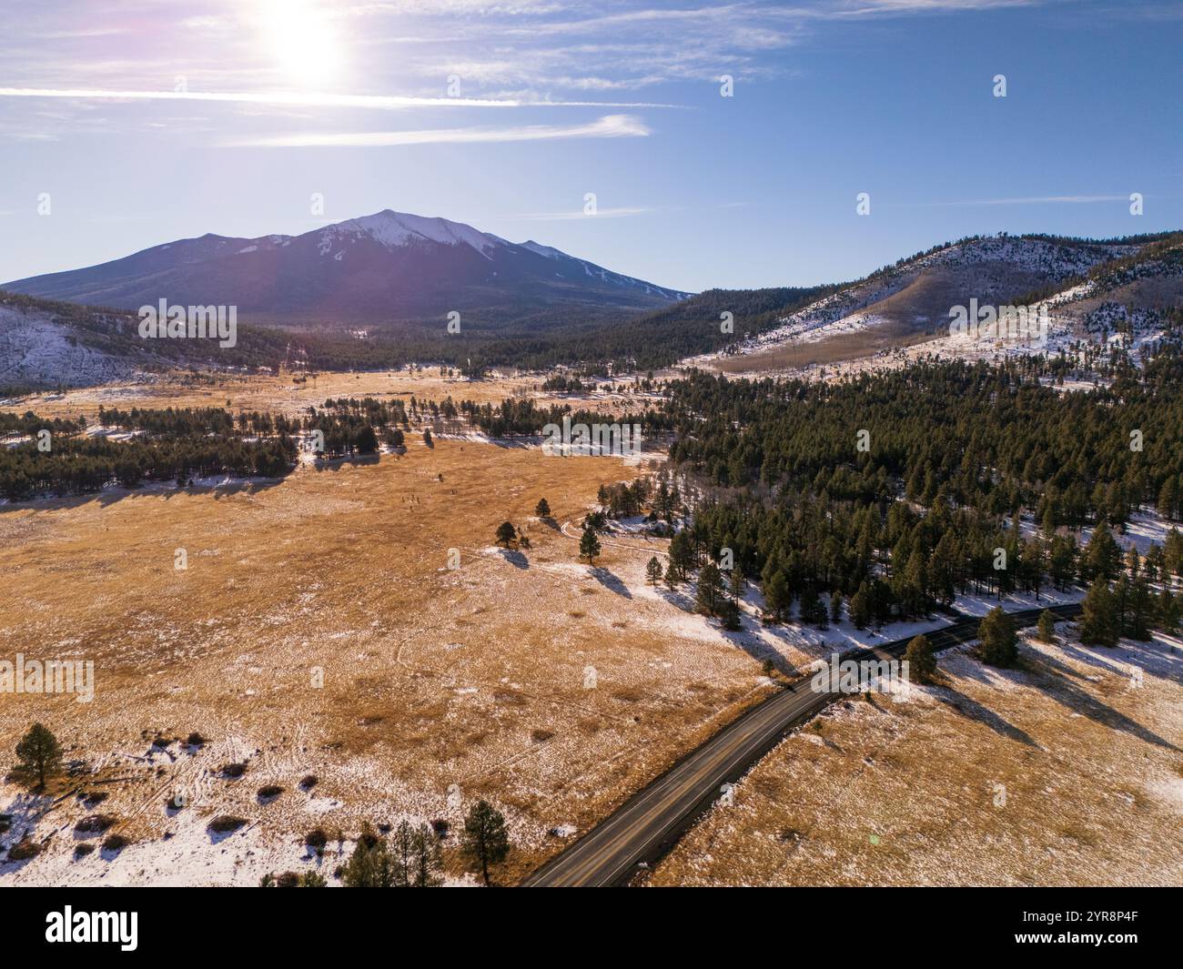 Vista aerea ad angolo elevato di una strada di montagna che attraversa un prato innevato e una foresta di pini verso il Humphreys Peak sotto un sole luminoso vicino a Flagstaff Foto Stock