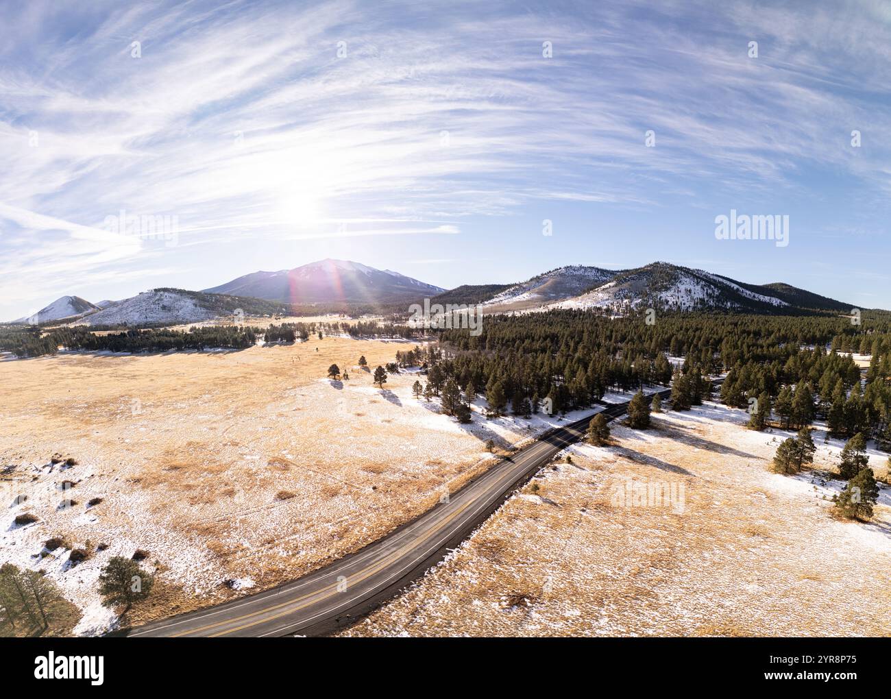Vista aerea ad angolo elevato di una strada di montagna che attraversa un prato innevato e una foresta di pini verso il Humphreys Peak sotto un sole luminoso vicino a Flagstaff Foto Stock