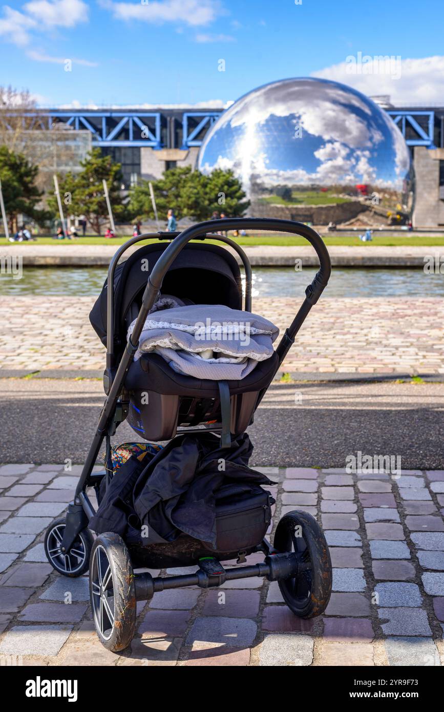 Il bambino dorme in un passeggino in una giornata di sole con la cupola de la Géode sullo sfondo, lungo il Canal de l'Ourcq a Parc de la Villette, Parigi, Francia. Foto Stock