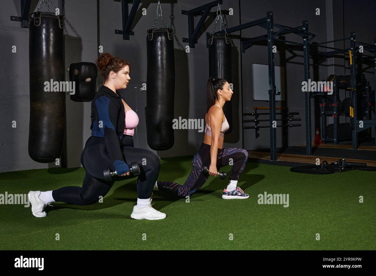 Due donne eseguono affondamenti con pesi in un ambiente di palestra spazioso e ben attrezzato. Foto Stock Due donne eseguono affondamenti con pesi in un ambiente di palestra spazioso e ben attrezzato. Foto Stock