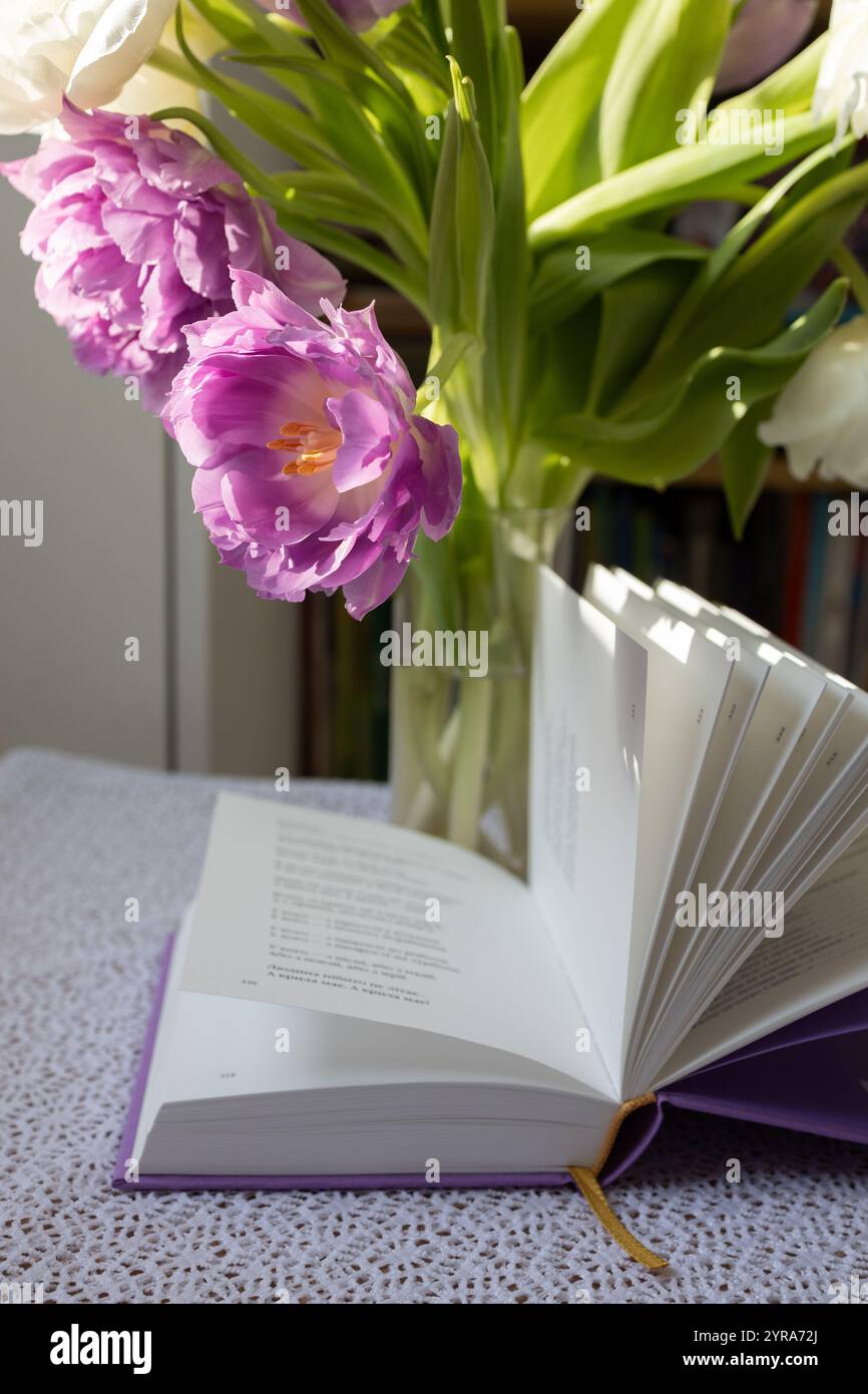 un libro aperto con poesie, fiori di tulipano nelle vicinanze. Mattinata accogliente, piacevole solitudine, giornata di sole. Semplice gioia umana. Hobby - leggere libri Foto Stock
