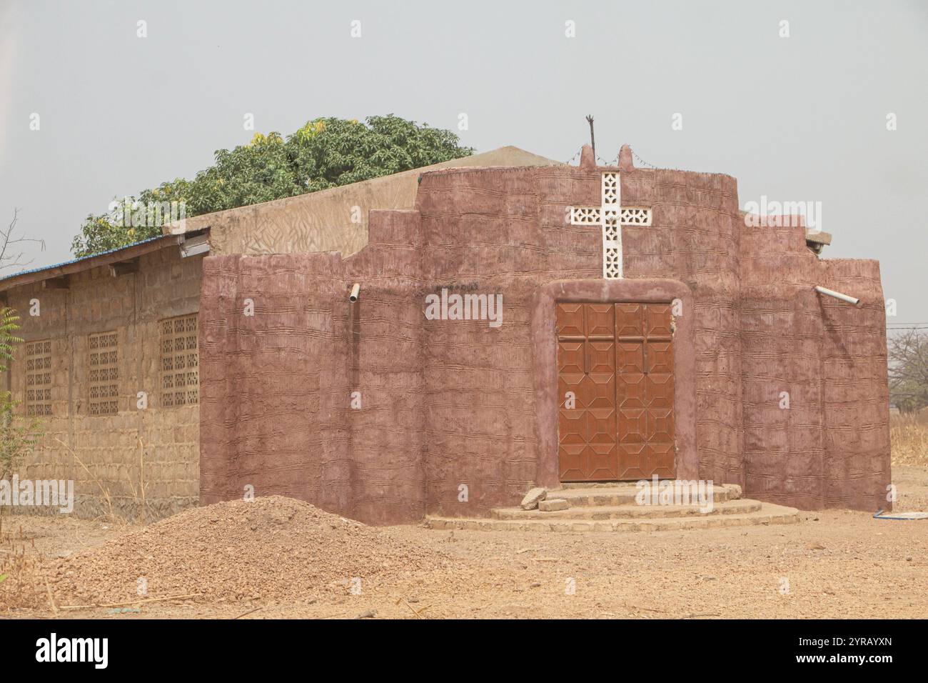 Chiesa rustica del Villaggio in argilla con una croce e porta in legno nel Togo rurale, circondata da paesaggio secco Foto Stock
