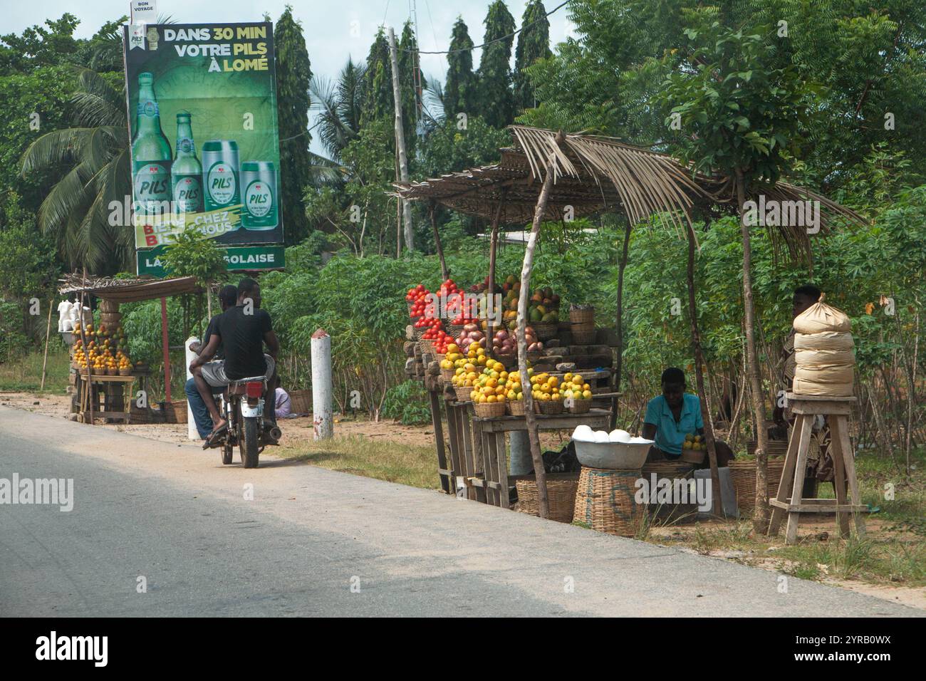 Bancarelle di frutta a bordo strada in Togo con prodotti freschi e un cartellone pubblicitario rustico sullo sfondo Foto Stock
