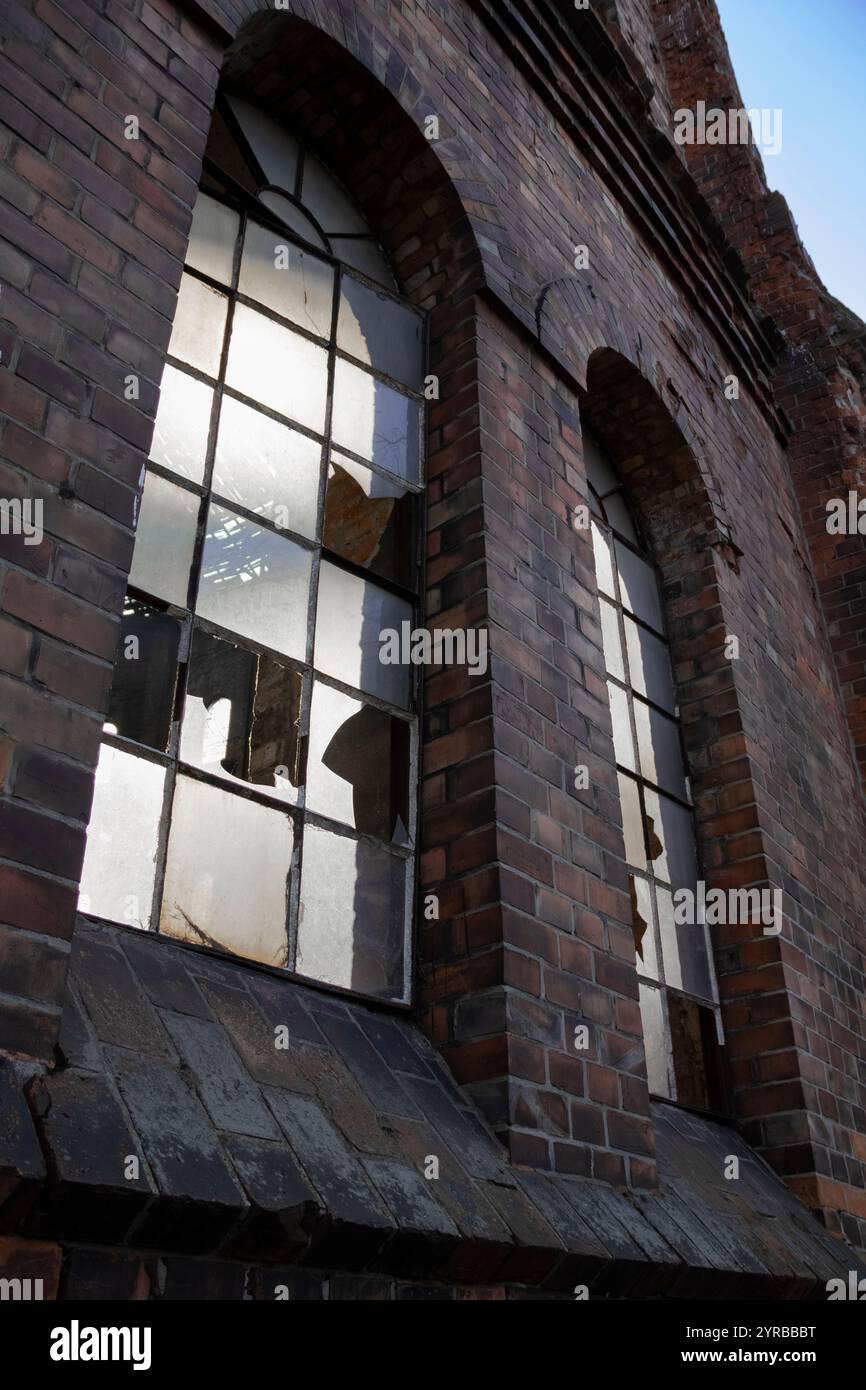 Un primo piano di vetro frantumato nelle finestre di un edificio industriale abbandonato in mattoni, adagiato su uno sfondo urbano grintoso. Foto Stock