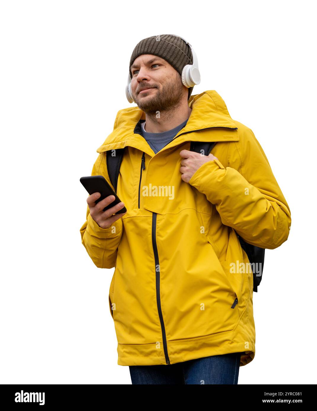 Uomo caucasico con giacca e cappello invernali gialli, in piedi con un telefono cellulare, cuffie wireless e uno zaino. Foto Stock