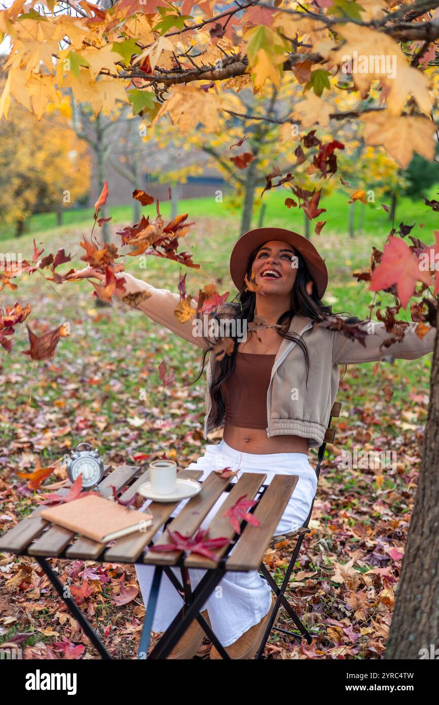 La giovane e allegra donna latina che indossa un cappello lancia foglie autunnali nell'aria mentre si siede a un tavolo di legno in un parco, si gusta una tazza di caffè e. Foto Stock