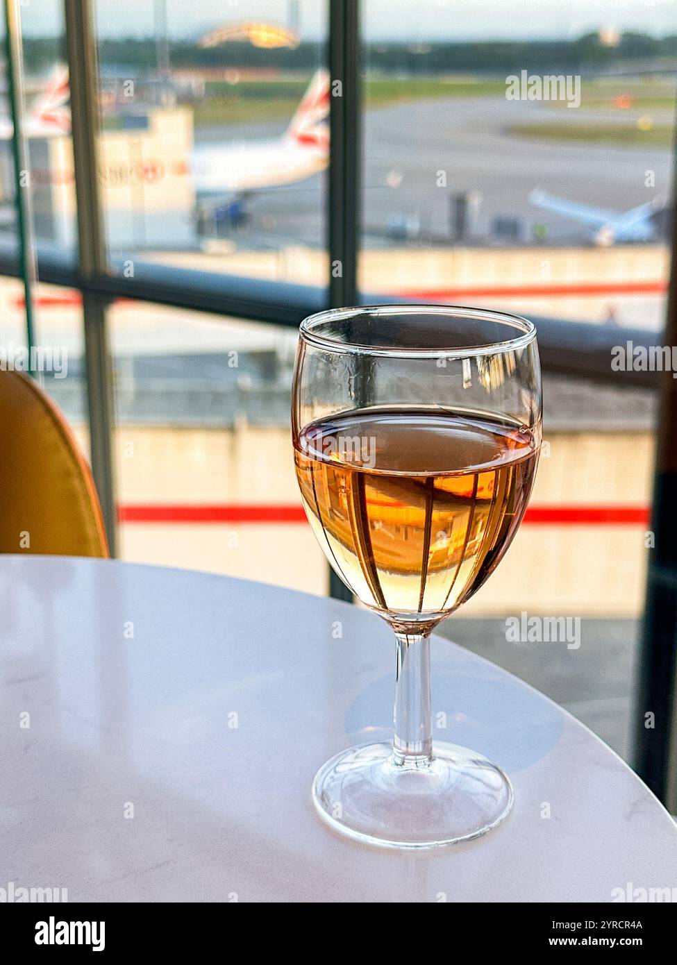 Londra, Inghilterra, Regno Unito - 21 agosto 2024: Bicchiere di vino rosa su un tavolo in una business lounge con vista sugli aerei della British Airways fuori dal Terminal 5 - Immagine stock catturata con smartphone