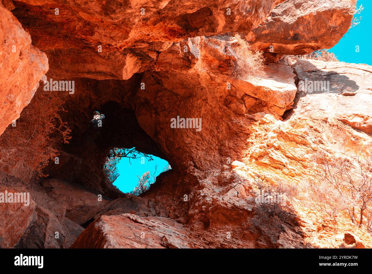 Una vibrante vista di un ingresso roccioso nella grotta illuminato dalla luce del sole, che mostra vivaci tonalità arancio e rosso con uno sguardo sul cielo blu luminoso della ba Foto Stock